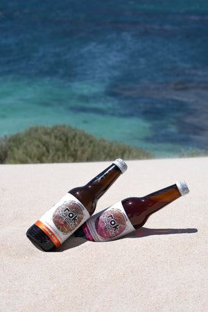Two bottles of rok Kombucha lying on white sand above a turquoise bay, Margaret River WA