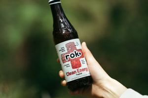Hand holding a bottle of rok+ Clean Energy Tropical kombucha energy drink outdoors